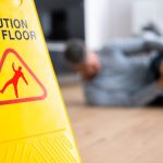 Close up of yellow Caution Wet Floor sign with man laying on floor in the background holding his knee