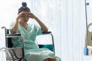 Woman in a wheelchair in a hospital room her elbows are on the arms of the chair and she is holding her head in her hands